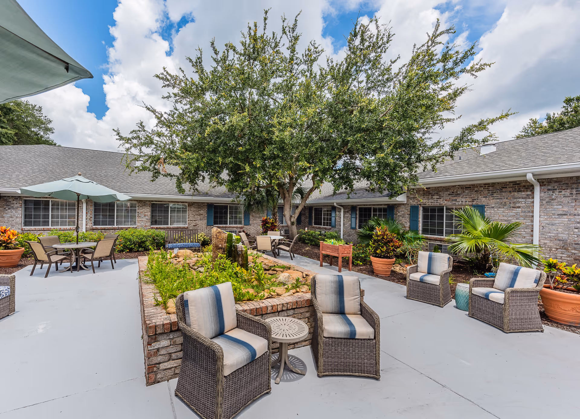Outdoor courtyard area with patio furniture including wicker chairs with cushions, a small table, and a table with an umbrella. There is a large tree in the center surrounded by a raised brick planter with greenery. The courtyard is enclosed by a single-story brick building with windows and blue shutters.
