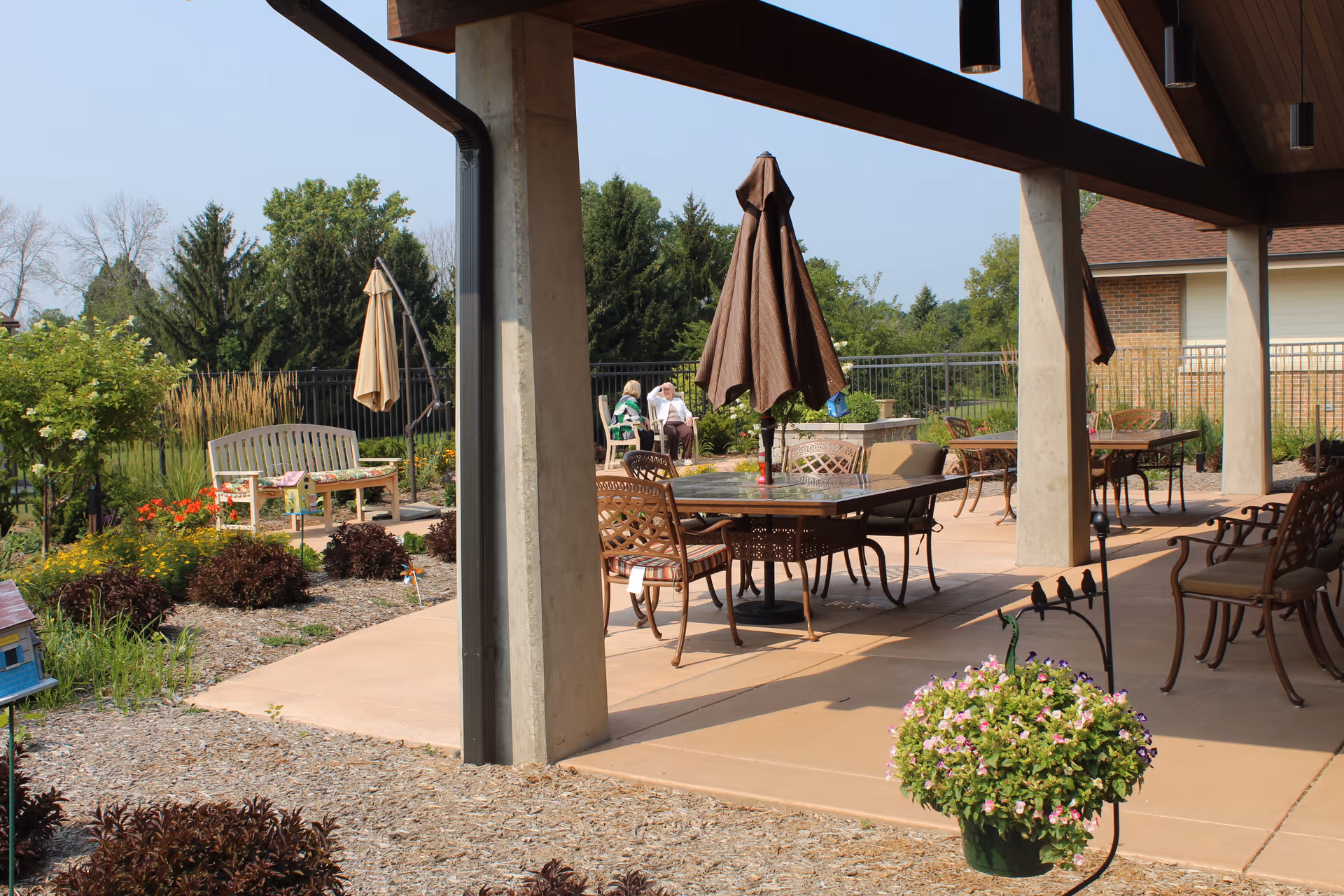 Covered outdoor patio and garden area with tables, chairs, umbrellas and benches at a senior living facility.
