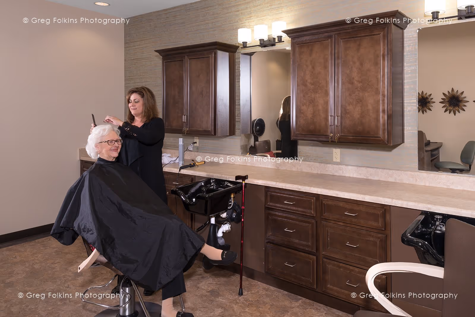 An elderly woman with white hair and glasses sits in a salon chair wearing a black cape while a hairstylist stands behind her, combing her hair. The salon area features dark wooden cabinets, a countertop, large mirrors, and a black hair washing sink. The setting appears clean and well-lit.