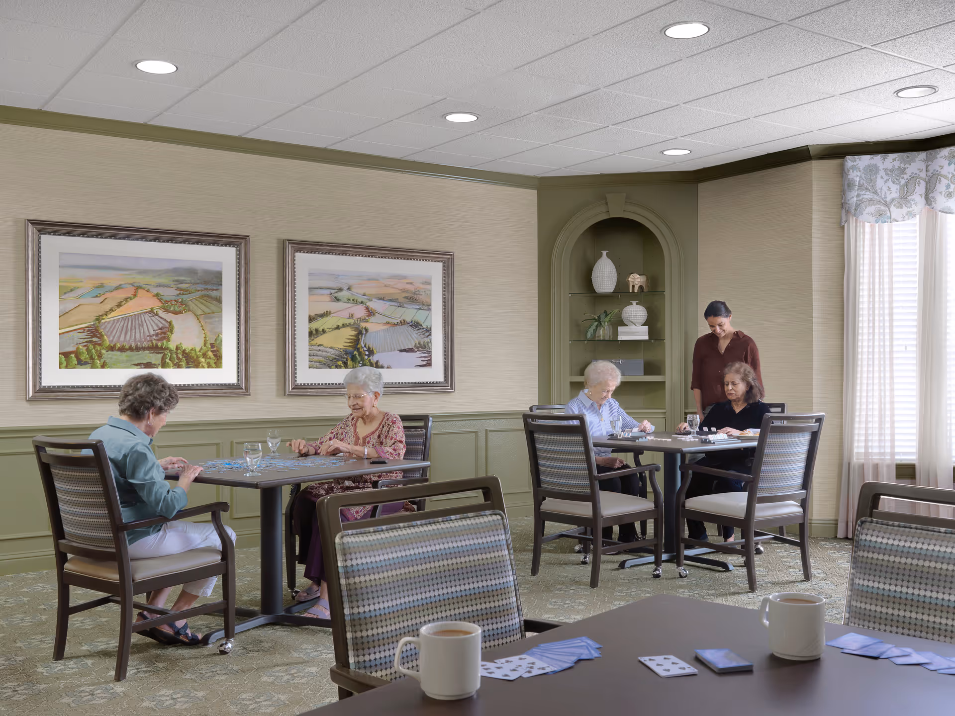 A cozy room with four elderly women sitting at two separate tables playing card and puzzle games. A younger woman stands nearby, smiling. The room has beige walls with green wainscoting, two framed landscape paintings, a built-in shelf with decorative items, and large windows with sheer curtains letting in natural light.