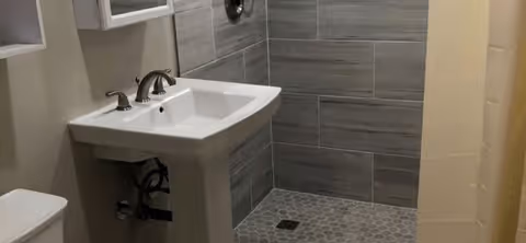 A bathroom featuring a white pedestal sink with silver faucets, a toilet to the left, and a walk-in shower with gray tiled walls and a hexagonal tiled floor. The shower has a drain in the center and a shower curtain on the right side.