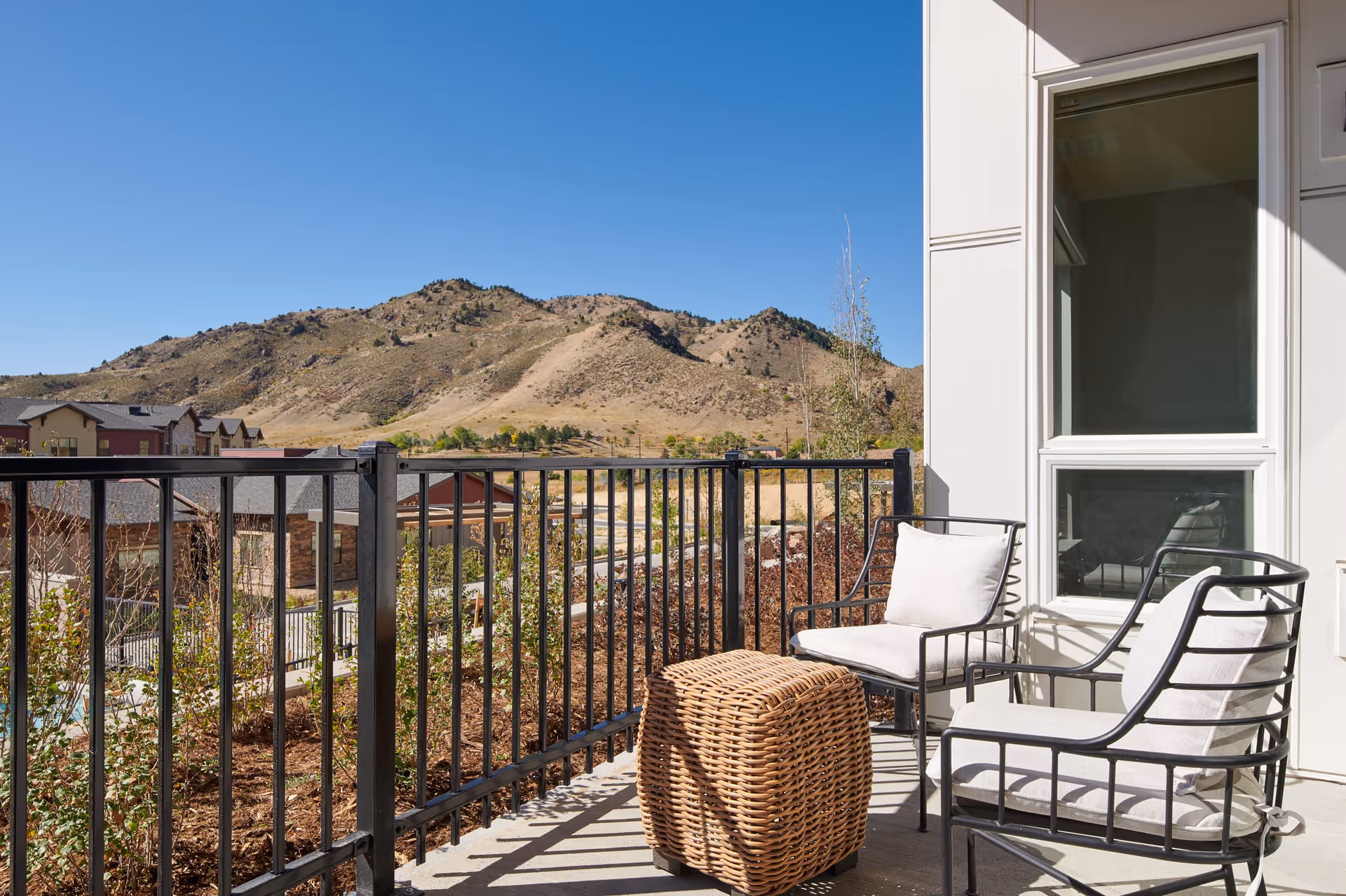 Outdoor patio area with two cushioned metal chairs and a woven rattan ottoman, overlooking a scenic view of dry hills and residential buildings under a clear blue sky.