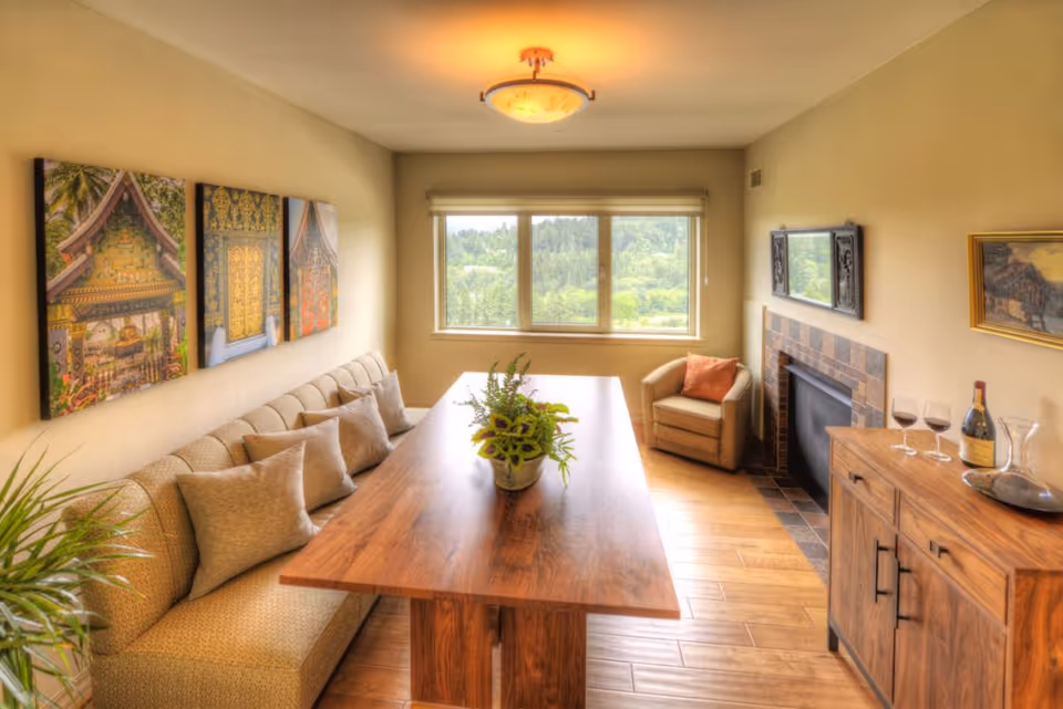 A cozy dining room with a long wooden table surrounded by a cushioned bench with multiple pillows on one side and a single armchair with an orange pillow near a window overlooking greenery. The room features a fireplace with a decorative mirror above it, a wooden sideboard with wine glasses, a bottle of wine, and a decanter. Three framed pictures hang on the wall above the bench, and the floor is wooden with a warm tone.
