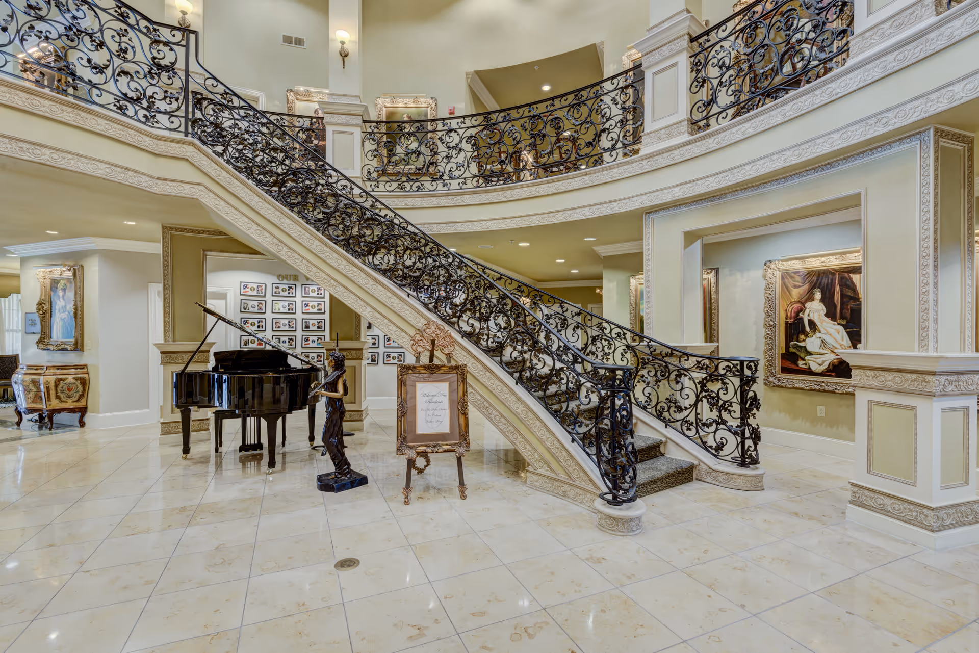 Elegant interior of a senior living facility featuring a grand curved staircase with ornate black wrought iron railings, a polished black grand piano, a bronze statue, framed artwork on the walls, and a decorative easel with a sign. The floor is tiled with light-colored marble, and the space is well-lit with wall sconces and recessed lighting.