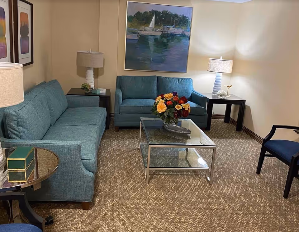 A cozy living room featuring a teal sofa and armchair, a glass coffee table with a floral arrangement, and decorative lamps in a warm, inviting setting.