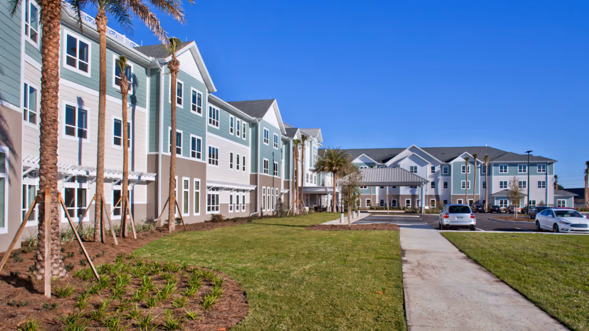Exterior view of a senior living facility named Trinity Springs with a well-maintained lawn, palm trees, a paved walkway, and parked cars under a clear blue sky.