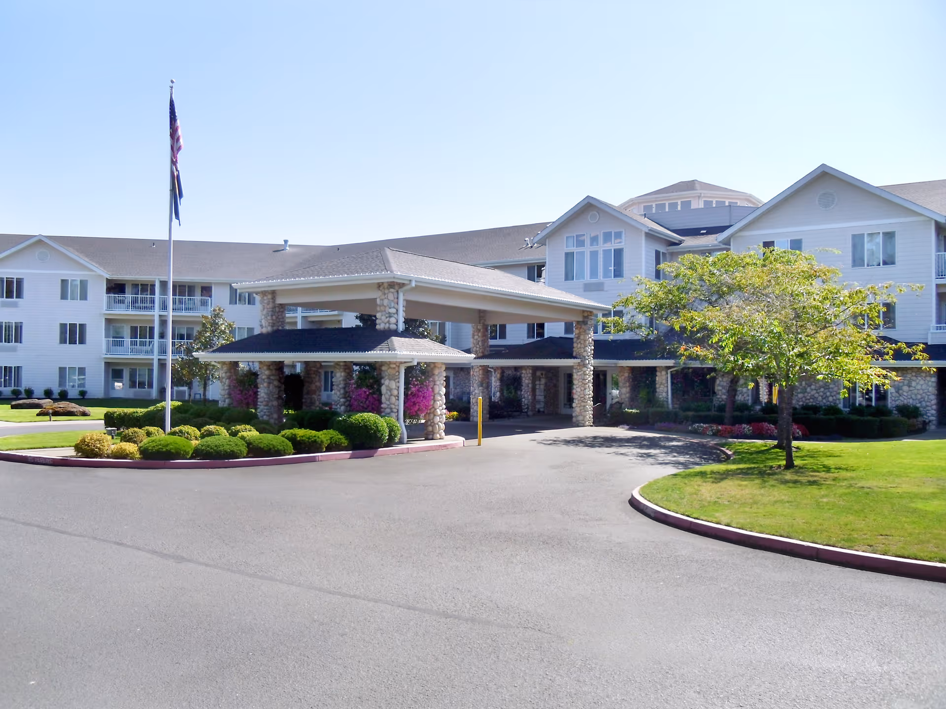 Exterior view of Somerset Lodge Gracious Retirement Living, showing a large multi-story building with white siding and stone pillars at the entrance. There is a covered driveway with landscaping including bushes, flowers, and a tree, and two flagpoles with flags flying in front of the building under a clear blue sky.