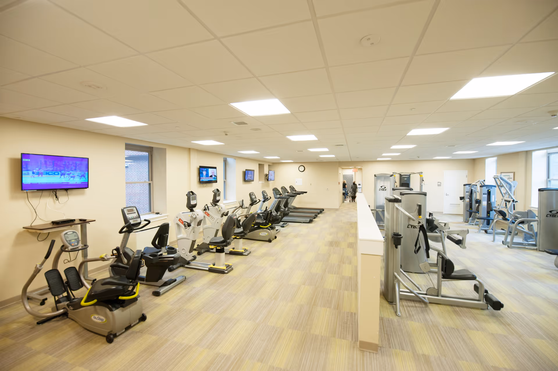 A spacious fitness room with various exercise equipment including stationary bikes, treadmills, and weight machines. The room has a light-colored floor and ceiling with multiple ceiling lights. Several wall-mounted TVs are visible, and there are windows letting in natural light.