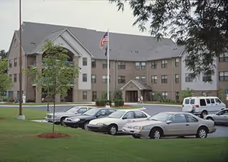 Exterior view of a three-story senior living facility building with a gray roof and beige walls. There is a flagpole with an American flag in front of the building, several parked cars in the parking lot, and green grass with a few trees around the area.