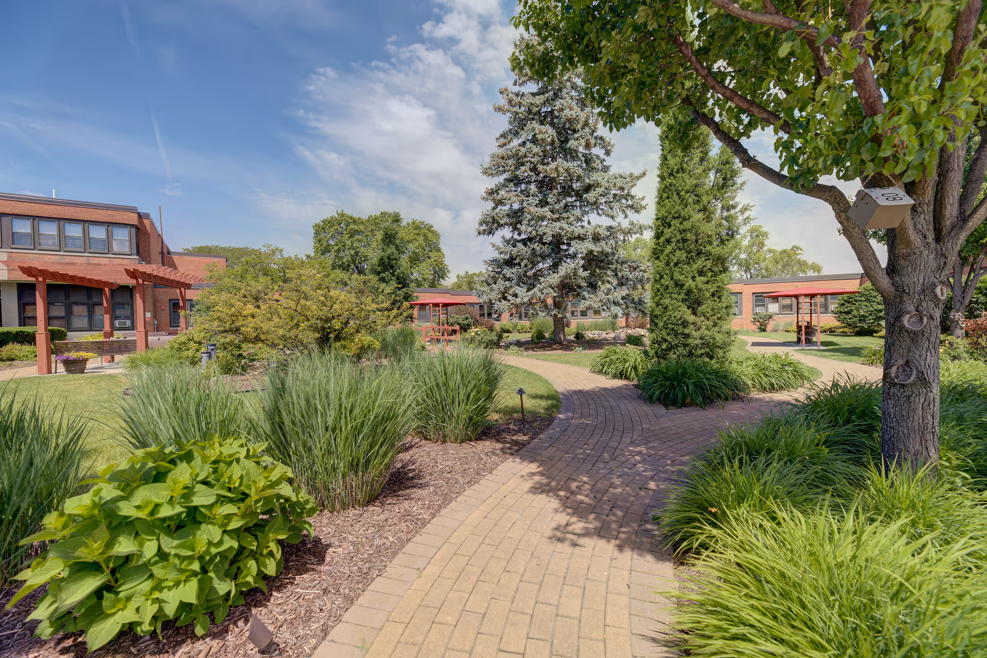 A landscaped outdoor garden area with a winding brick pathway surrounded by green shrubs, trees, and plants. In the background, there are single-story and two-story brick buildings with red awnings under a partly cloudy blue sky.