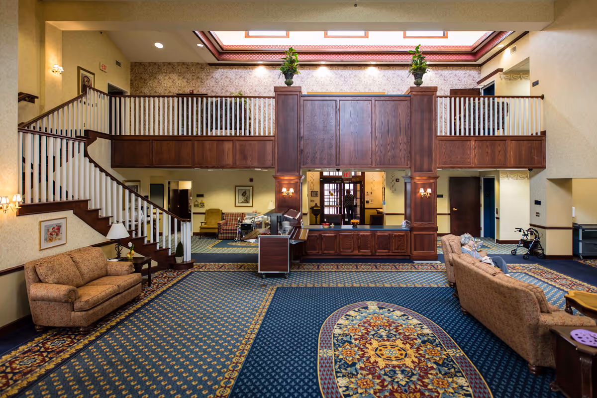 Spacious senior living facility common area with patterned carpet, floral wallpaper, and wooden railings on the upper level. There are multiple seating areas with sofas and chairs, a staircase on the left, and a reception or service counter in the center. Two elderly people are seated on a sofa on the right side, and a person is visible near the entrance in the background.