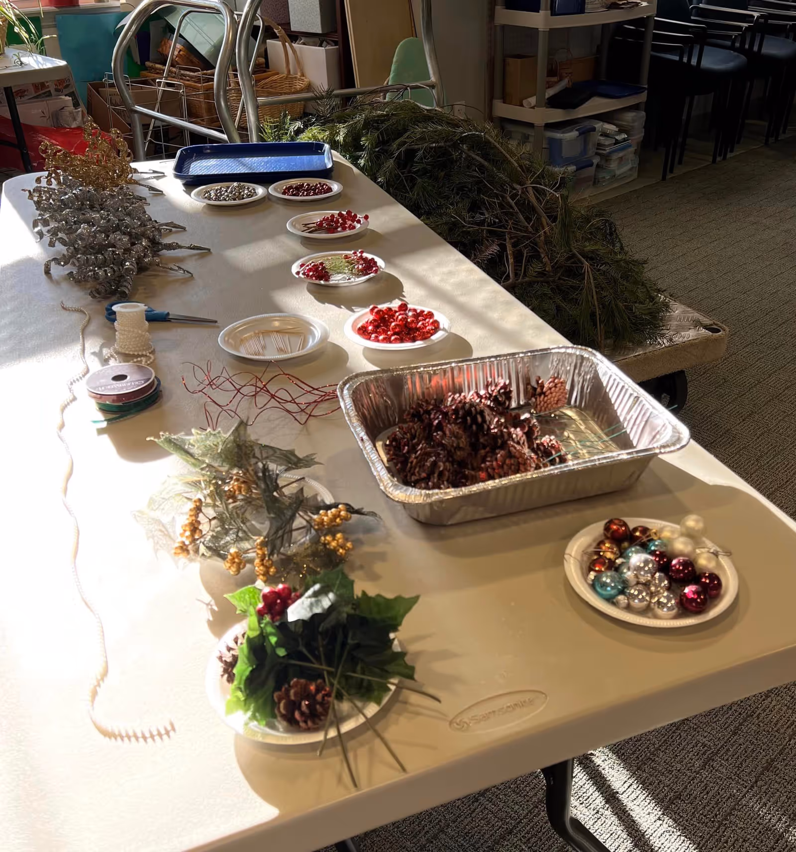 A table set up with holiday craft supplies including pinecones, ornaments, greenery, ribbons and trays in a communal room.