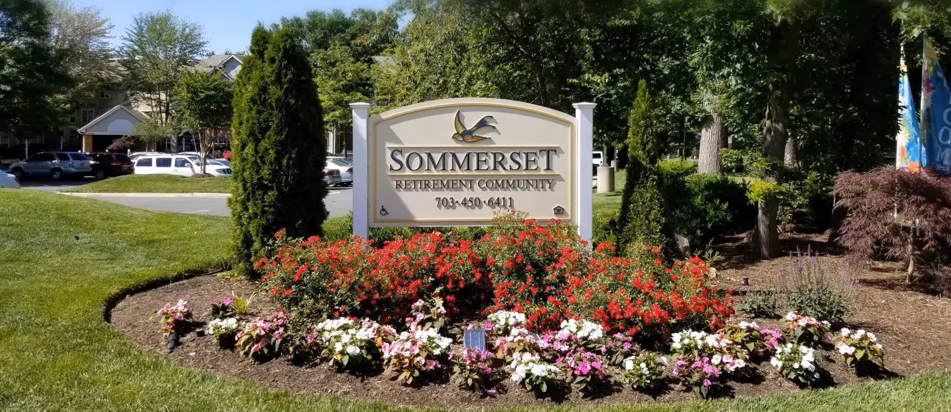 A landscaped entrance sign for Sommerset Retirement Community surrounded by colorful flowers and greenery, with trees and parked cars in the background under a clear blue sky.