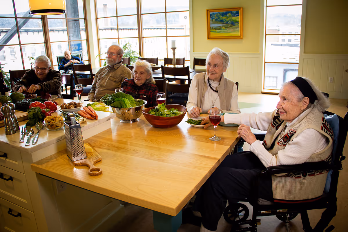 Several older adults sit around a communal dining table with bowls of salad and vegetables in a bright dining area.