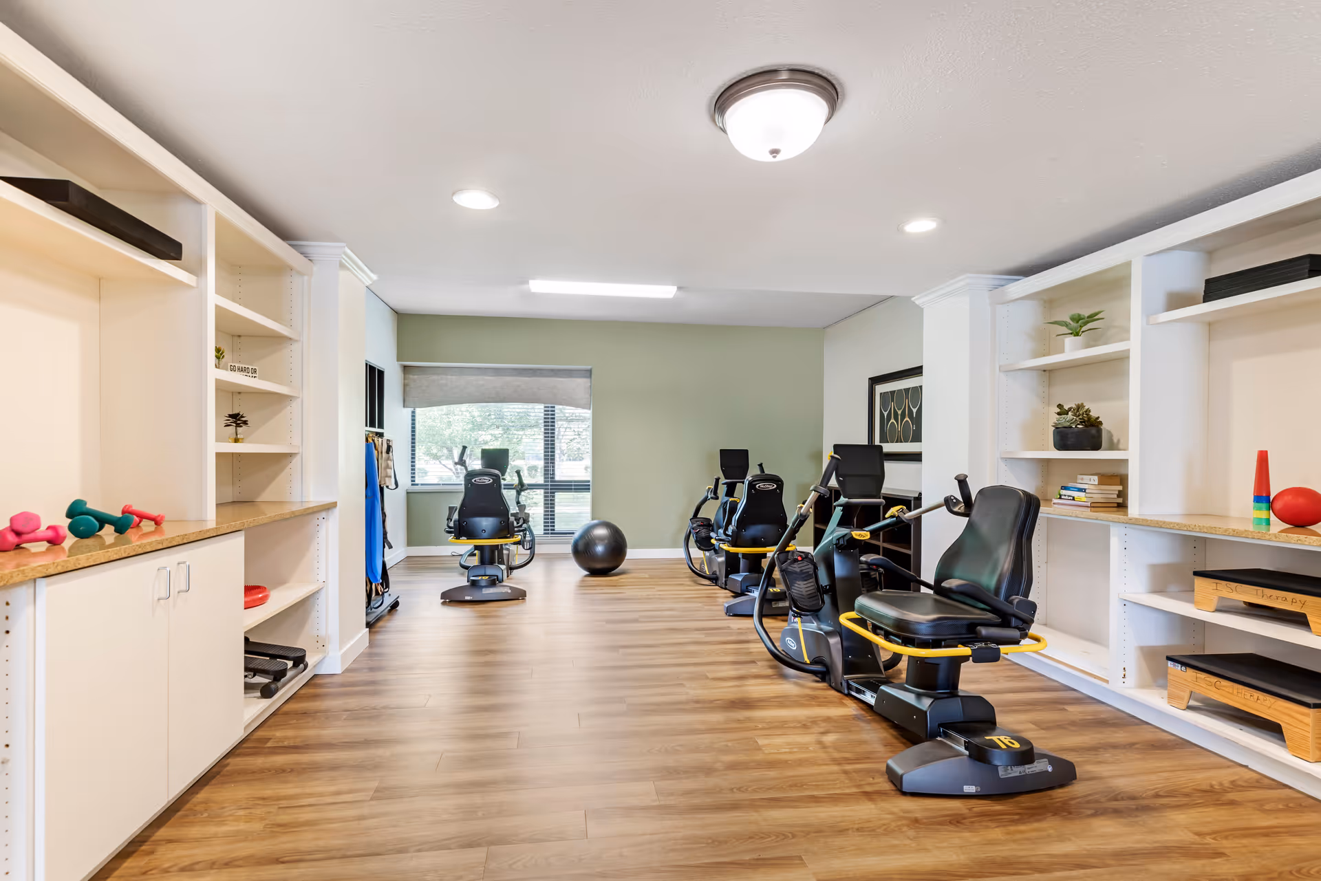 A fitness room with exercise equipment including recumbent bikes, a large exercise ball, and shelves with small weights, books, and decorative plants. The room has wood flooring, white walls with a green accent wall, and a window with blinds letting in natural light.