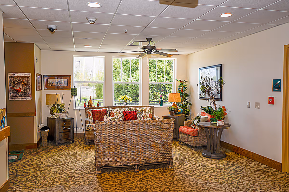A cozy living room area with wicker furniture including a sofa and armchair adorned with floral and red cushions. The room has large windows letting in natural light, a ceiling fan, several potted plants, framed artwork on the walls, and a patterned carpet.
