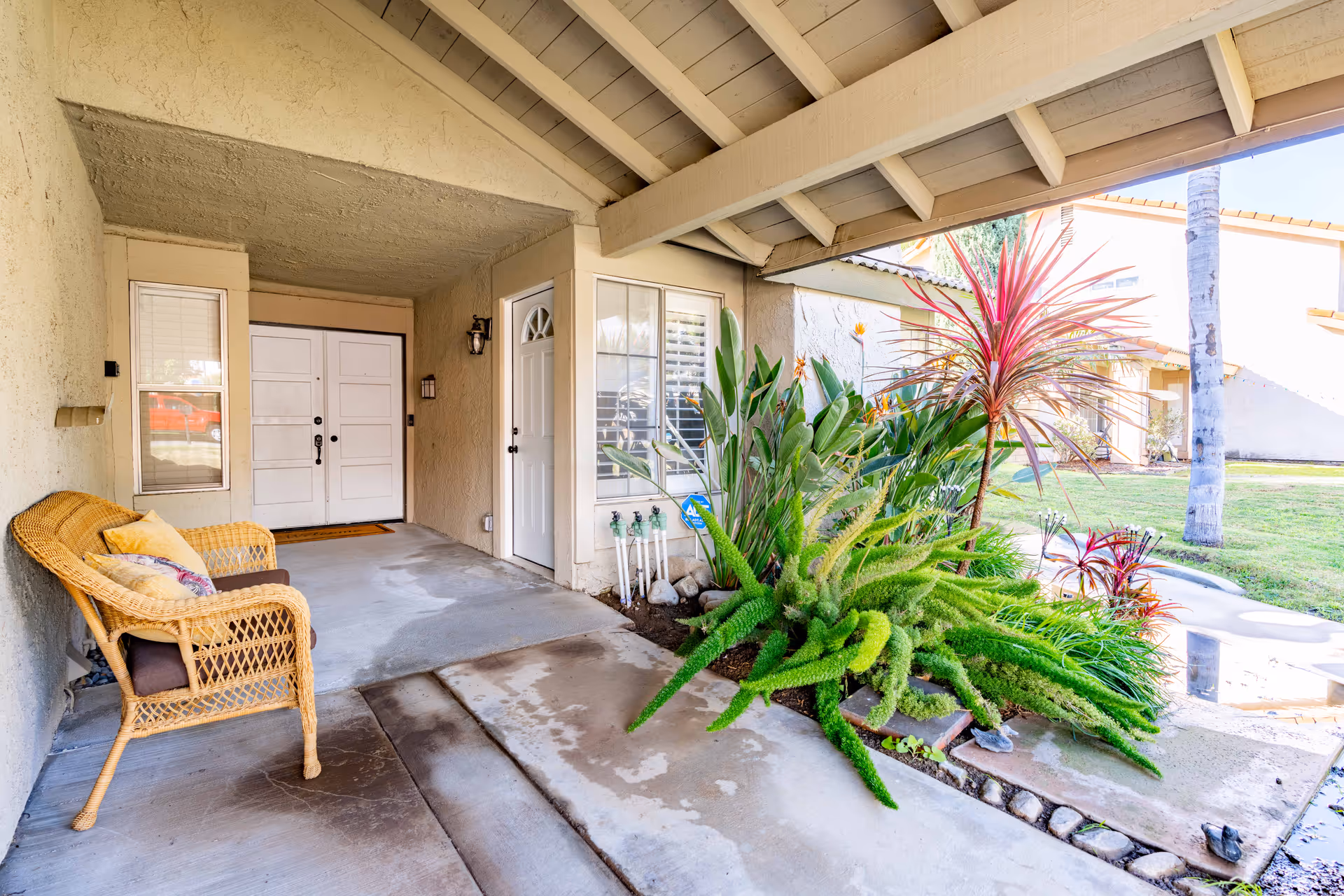 Covered entrance area of a building with a white double door and a single white door, a wicker bench with cushions on the left, and a garden bed with various green and red plants on the right. The area is partially shaded by a wooden roof structure, and a grassy lawn with palm trees is visible in the background.