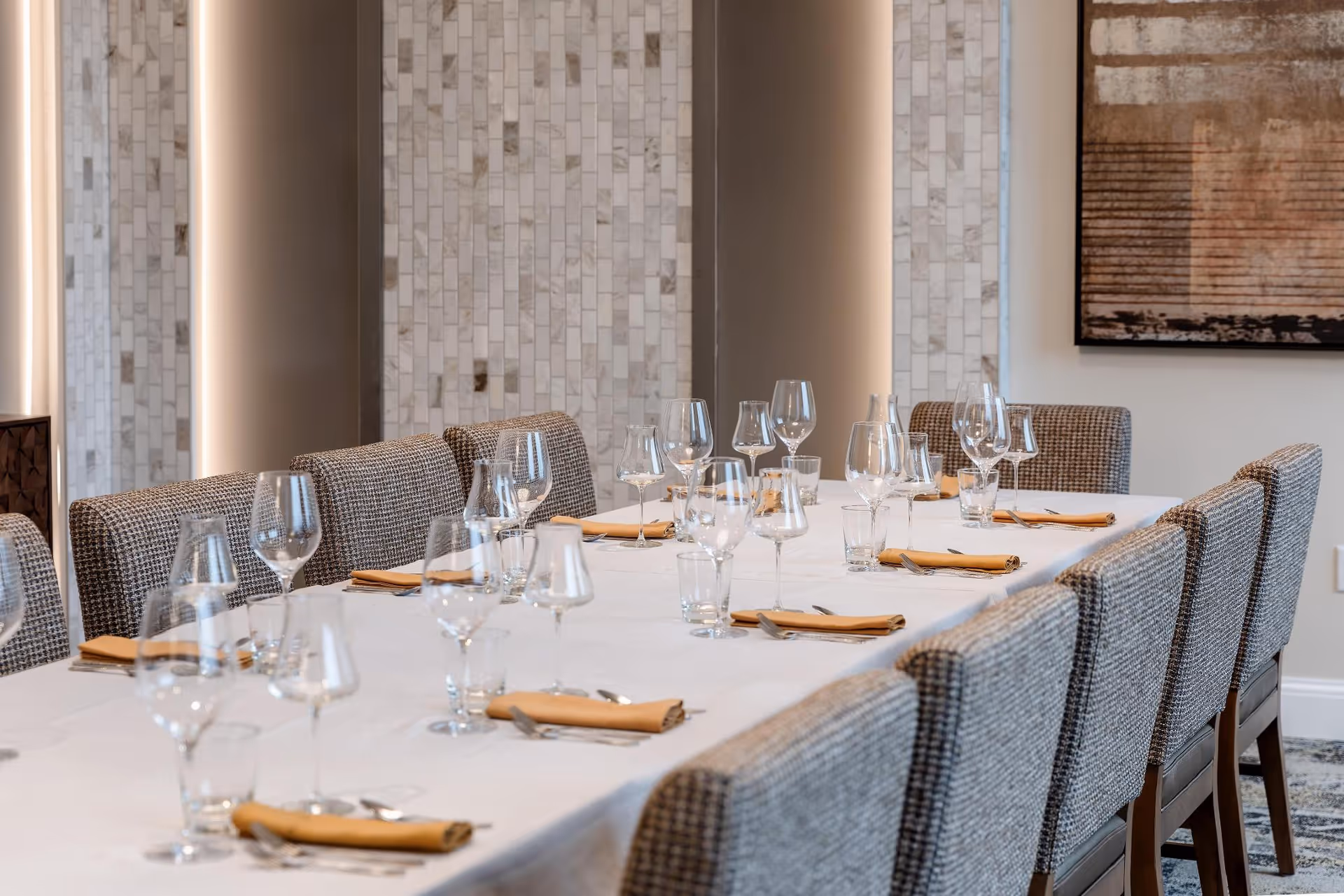 A long dining table set with clear wine glasses, water glasses, and folded yellow napkins on a white tablecloth. The table is surrounded by cushioned chairs with a textured fabric. The background features a tiled wall with vertical lighting and a framed abstract artwork on the right.