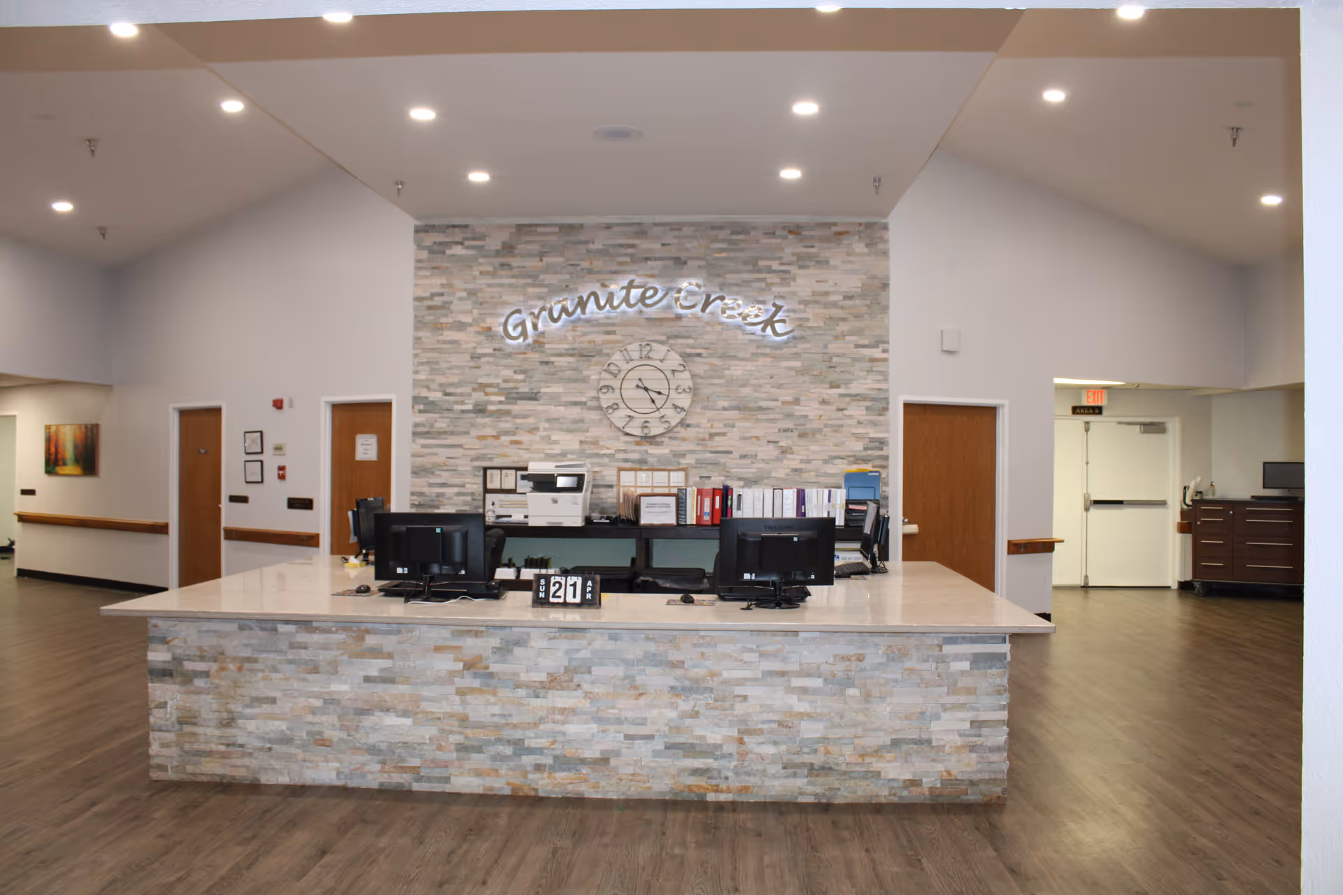 Reception desk area in Granite Creek Health and Rehabilitation facility with a stone accent wall behind it, featuring a clock and the facility name in illuminated letters. The desk has two computer monitors and office supplies, with doors and hallways visible in the background.