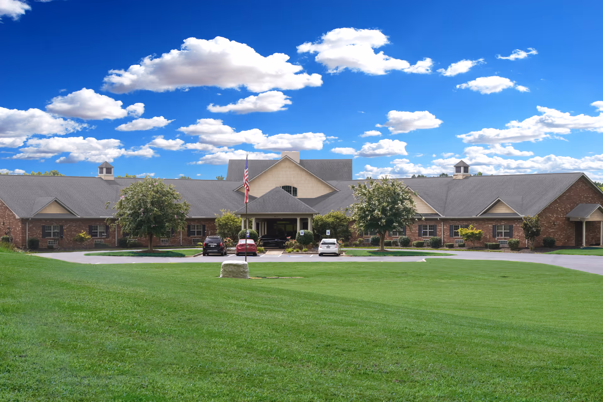 Front exterior view of Sunny Grove Assisted Living facility with a large green lawn in the foreground, a driveway with parked cars, an American flag on a flagpole, and a partly cloudy blue sky above.