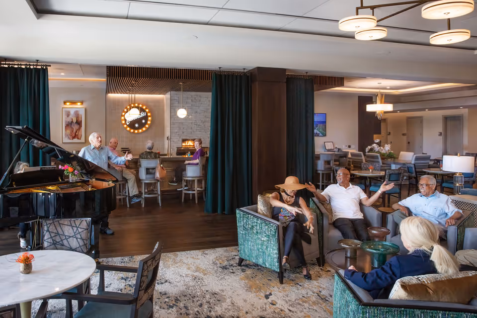A spacious and well-lit common area in a senior living facility with several elderly people sitting and conversing. The room features comfortable armchairs, a grand piano, a round table with chairs, and a bar area with stools. The decor includes modern lighting fixtures, dark curtains, and a patterned rug on the floor.
