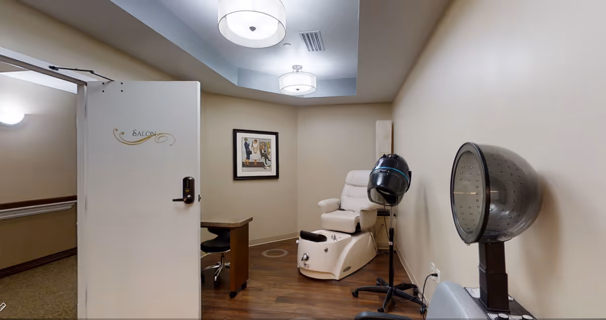 Interior view of a salon room with a white pedicure chair, a black salon hair dryer on a stand, a small desk with a black swivel chair, and a framed picture on the beige wall. The door to the room is open and labeled 'Salon'.