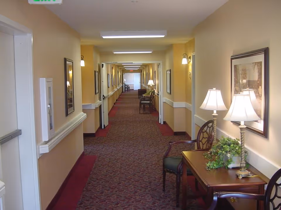 A long, well-lit hallway in an assisted living facility with beige walls and patterned carpet. The hallway features framed artwork on the walls, wall-mounted lights, and a small table with two lamps and a plant on the right side. Chairs are placed along the hallway for seating.