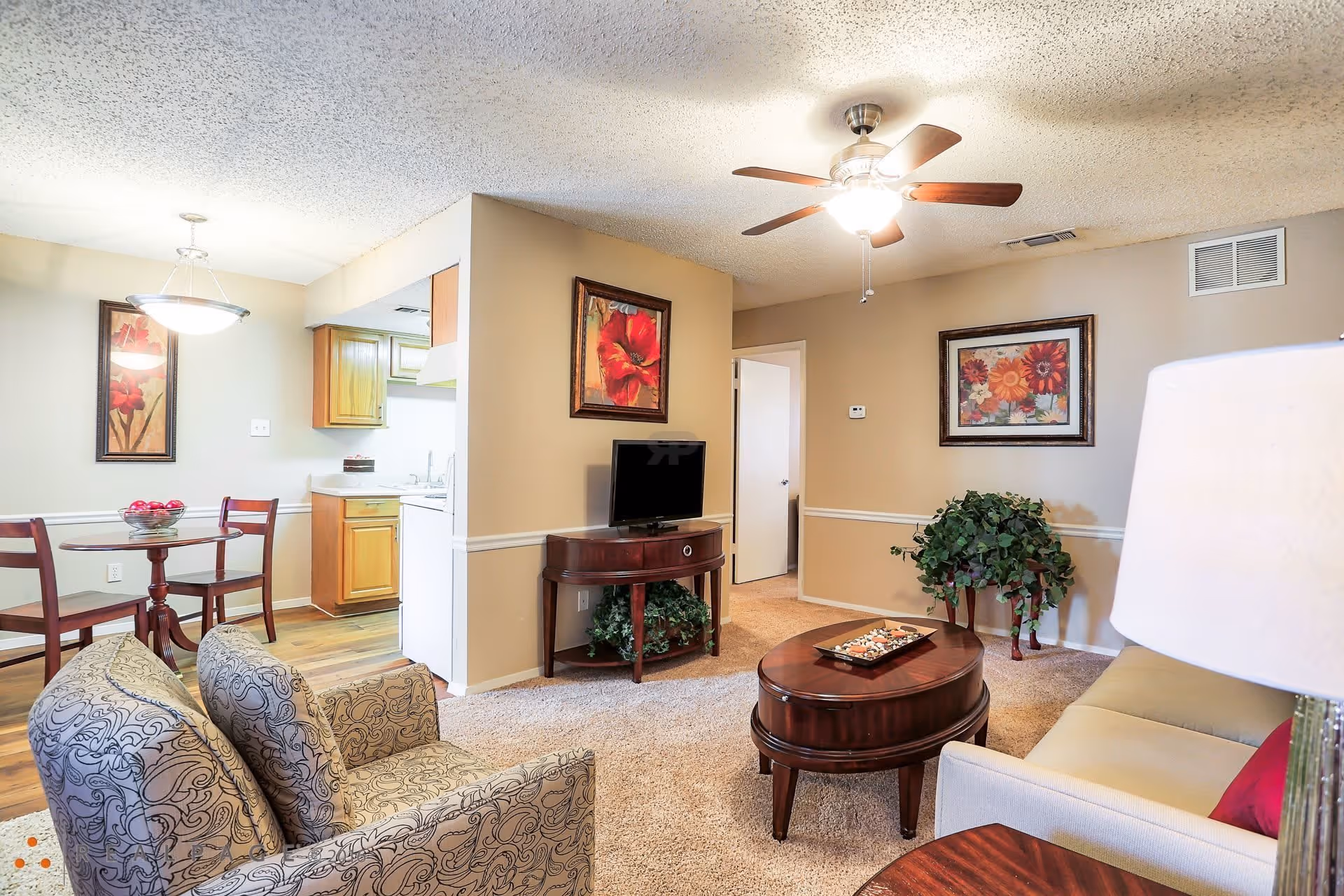 A cozy living room and dining area in MontClair Estates featuring a ceiling fan with light, a patterned armchair, a beige sofa with a red pillow, a wooden coffee table with decorative items, a small TV on a wooden stand, framed floral artwork on the walls, a dining table with two chairs and a bowl of red apples, and a partial view of a kitchen with wooden cabinets.