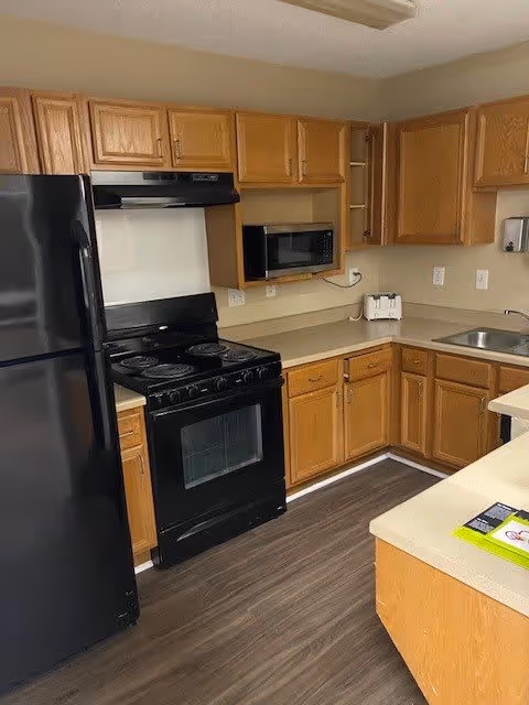 A kitchen area with wooden cabinets, a black refrigerator, a black stove with an oven, a microwave mounted above the counter, a toaster, a sink, and beige countertops. The floor is covered with dark wood-style flooring.
