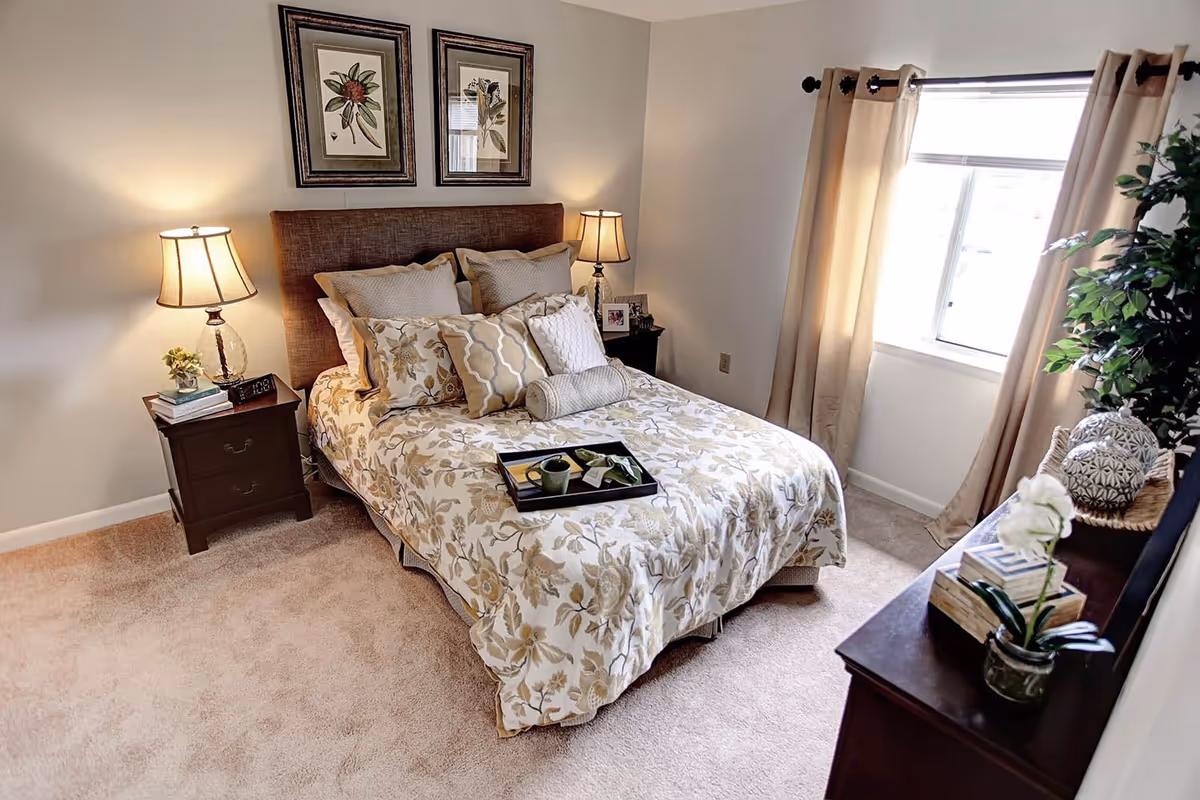 A cozy bedroom with a bed covered in a floral patterned comforter and multiple decorative pillows. Two bedside tables each hold a lamp and small decor items. A window with beige curtains allows natural light into the room. A dresser with decorative items and a potted plant is visible in the foreground.