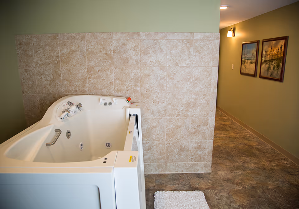 A walk-in bathtub with control knobs and a safety handle, positioned next to a tiled wall in a room with green walls and a tiled floor. A white bath mat is placed on the floor in front of the tub, and a hallway with framed pictures on the wall is visible in the background.