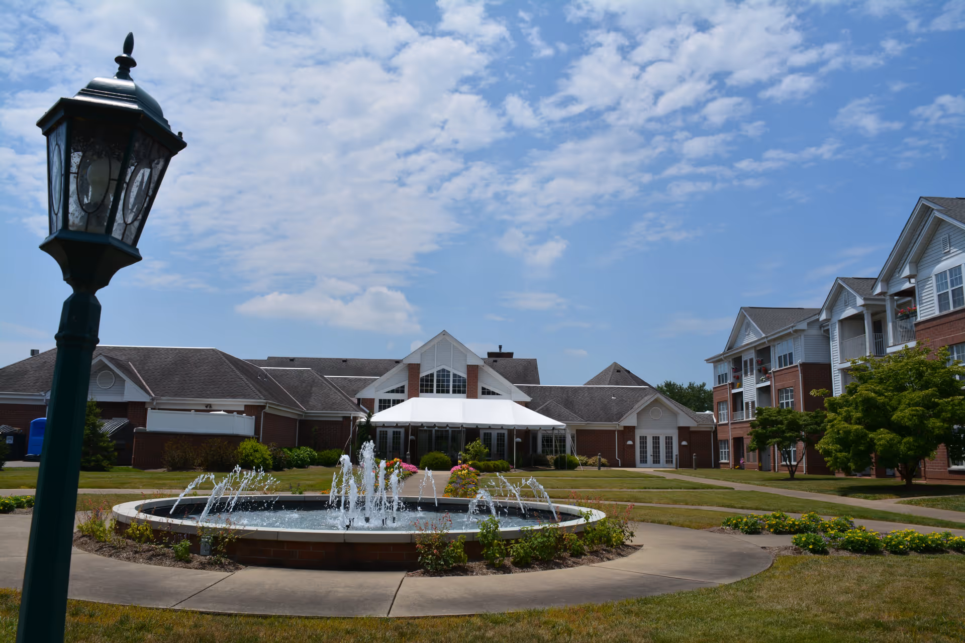 Outdoor courtyard area of The Glenwood Community with a circular water fountain in the center, surrounded by a paved walkway, green grass, and landscaped bushes. The background shows multi-story residential buildings with balconies and a partly cloudy blue sky.