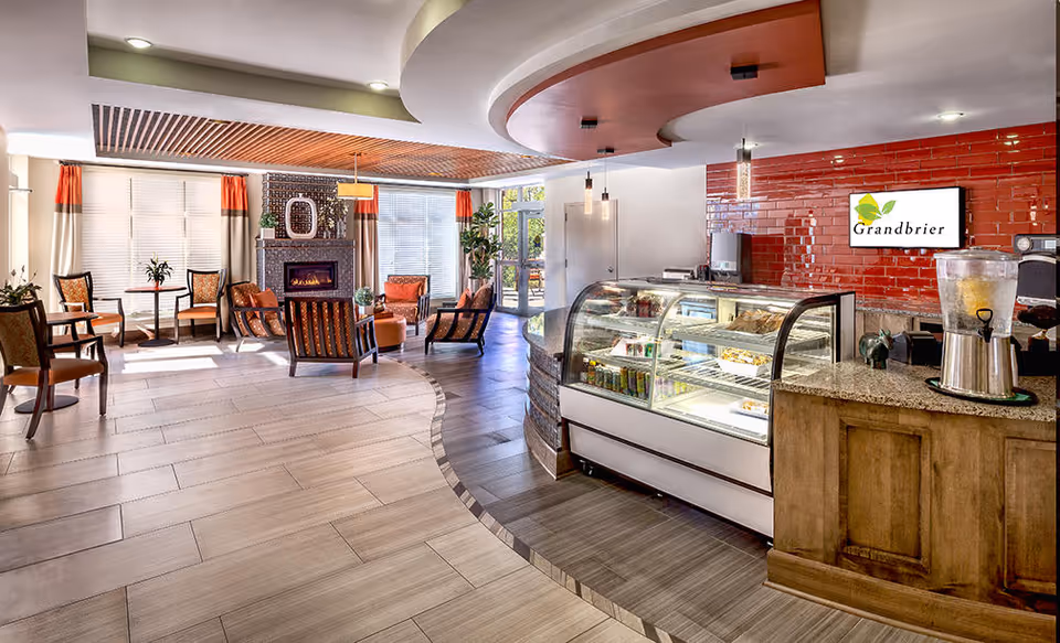 Bright senior living common area with seating around a fireplace and a café counter and pastry display on the right.