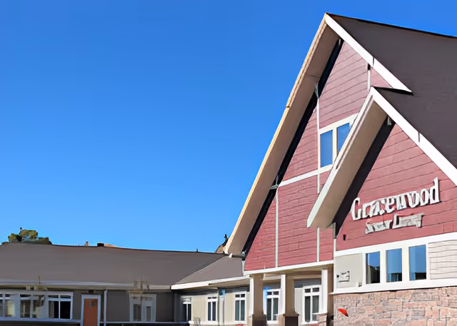 Exterior view of Gracewood Advanced Assisted Living and Memory Care building with a clear blue sky in the background. The building features a red and beige facade with multiple windows and a peaked roof.