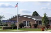 Exterior view of Gilman Healthcare Center, a single-story brick building with a flagpole flying the American flag in front. There are shrubs and potted plants near the entrance under a blue sign displaying the facility's name.
