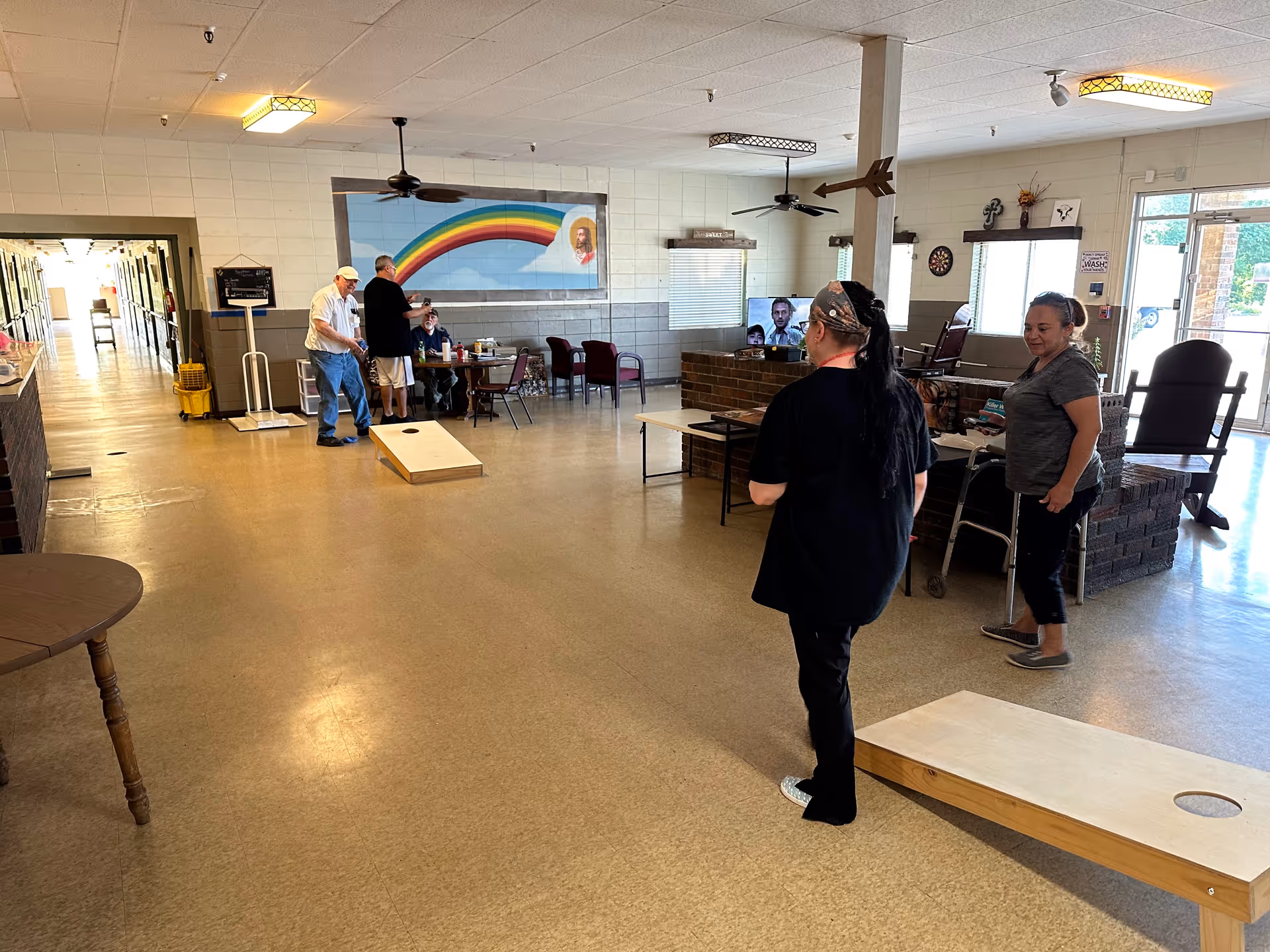 A spacious common area in a care center with several people engaging in a game of cornhole. The room has a tiled floor, ceiling fans, and fluorescent lighting. There is a large mural of a rainbow on the wall, a television, and various chairs and tables. A hallway extends from the left side of the room, and an exit door is visible on the right.
