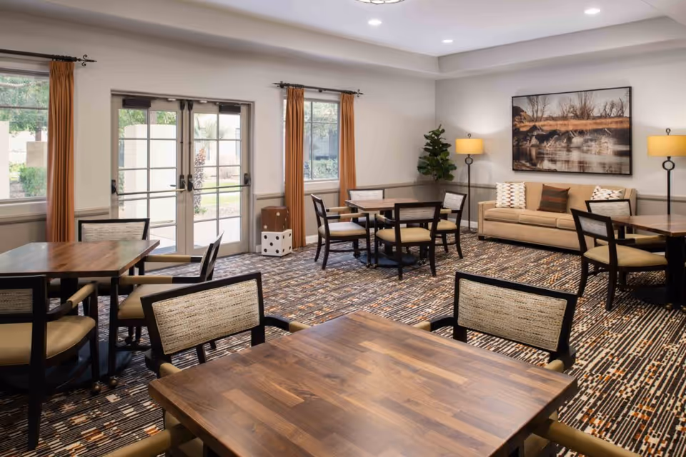 A well-lit common area with multiple wooden tables and cushioned chairs arranged on a patterned carpet. The room features large windows with orange curtains, a beige sofa with decorative pillows, two floor lamps, a large framed nature photograph on the wall, and a potted plant in the corner. Double glass doors lead outside to a garden area.