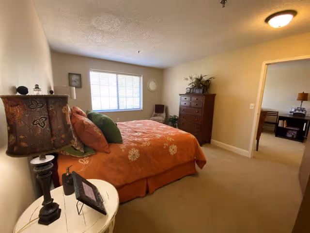 A bedroom with a large bed covered in an orange floral bedspread and multiple pillows. There is a bedside table with a lamp and picture frame, a tall wooden dresser with plants and decor on top, a window with blinds letting in natural light, and an open doorway leading to another room.
