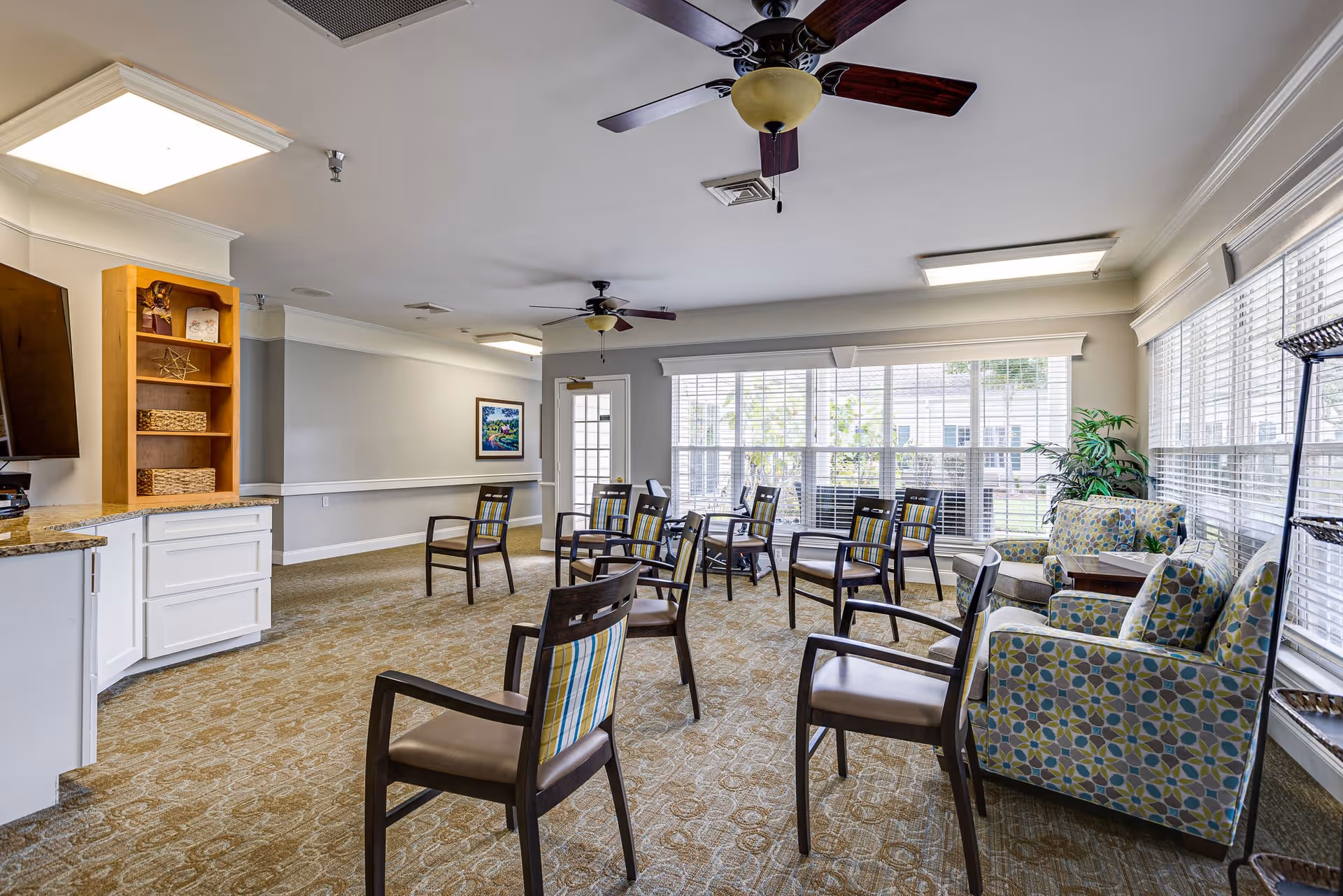 Sunlit senior living common room with chairs arranged in a circle, patterned armchairs by large windows, ceiling fans, and a small kitchenette counter.