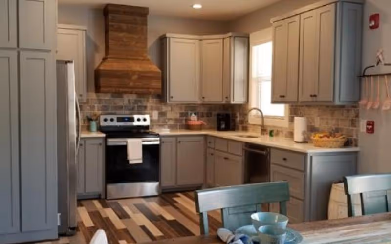 A modern kitchen with light gray cabinets, a stainless steel electric stove with a wooden range hood, a dishwasher, and a window above the sink. The kitchen has a brick-style backsplash and a wooden floor with varied tones. In the foreground, there is a wooden dining table with blue chairs and some tableware.