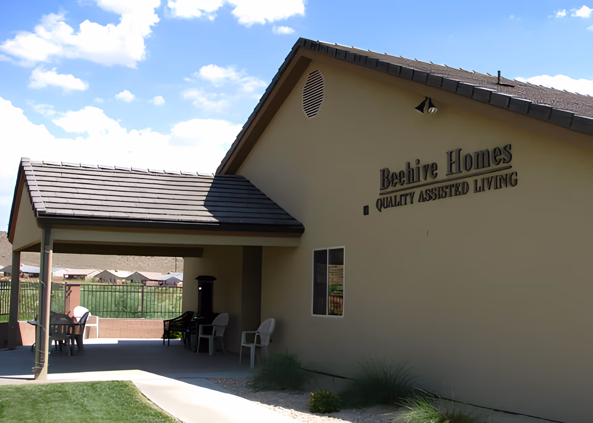 Exterior view of Beehive Homes assisted living facility showing a beige building with a covered patio area furnished with chairs and tables. The building has a tiled roof and a sign that reads 'Beehive Homes Quality Assisted Living'. The sky is blue with some clouds.