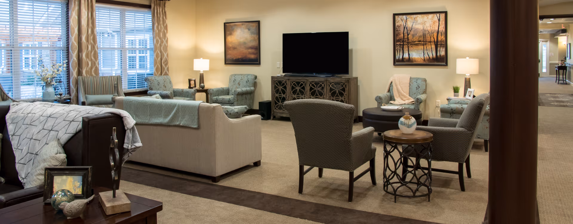 A cozy living room area in a senior living facility with multiple upholstered chairs and sofas arranged around a large flat-screen TV on a wooden cabinet. The room is warmly lit with table lamps and decorated with framed paintings on the walls, patterned curtains on the windows, and various decorative items on tables.