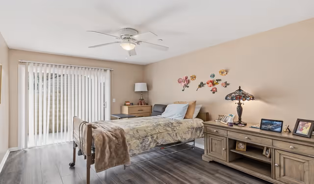 A senior living bedroom with a single bed covered in a floral blanket and a brown throw. The room has wood flooring, beige walls, and a ceiling fan with a light. There is a sliding glass door with vertical blinds leading outside. Next to the bed is a wooden nightstand with a lamp and some items on it. On the opposite side, there is a wooden dresser with a decorative lamp, framed photos, and small decor items. Colorful flower wall decorations are mounted above the bed.