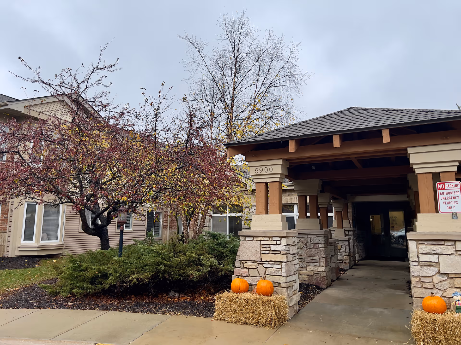 Entrance to a building with stone and wood pillars, a covered walkway, and the number 5900 displayed. There are two hay bales with pumpkins placed on them near the entrance. Trees with autumn leaves and bushes are visible on the left side under a cloudy sky.