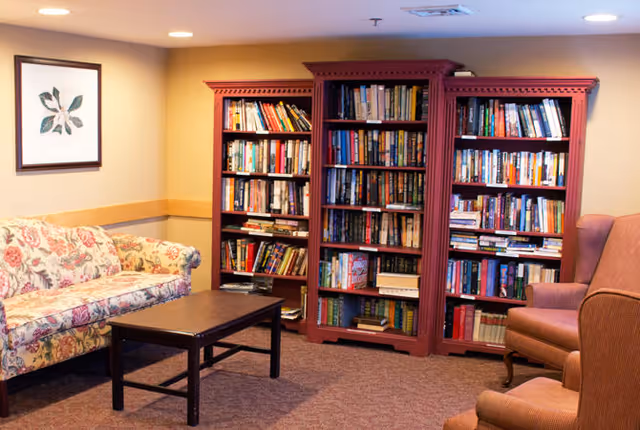 Cozy interior room with a floral patterned sofa, a dark wooden coffee table, two upholstered armchairs, and three large red bookshelves filled with books against the wall. A framed botanical print hangs on the wall above the sofa.
