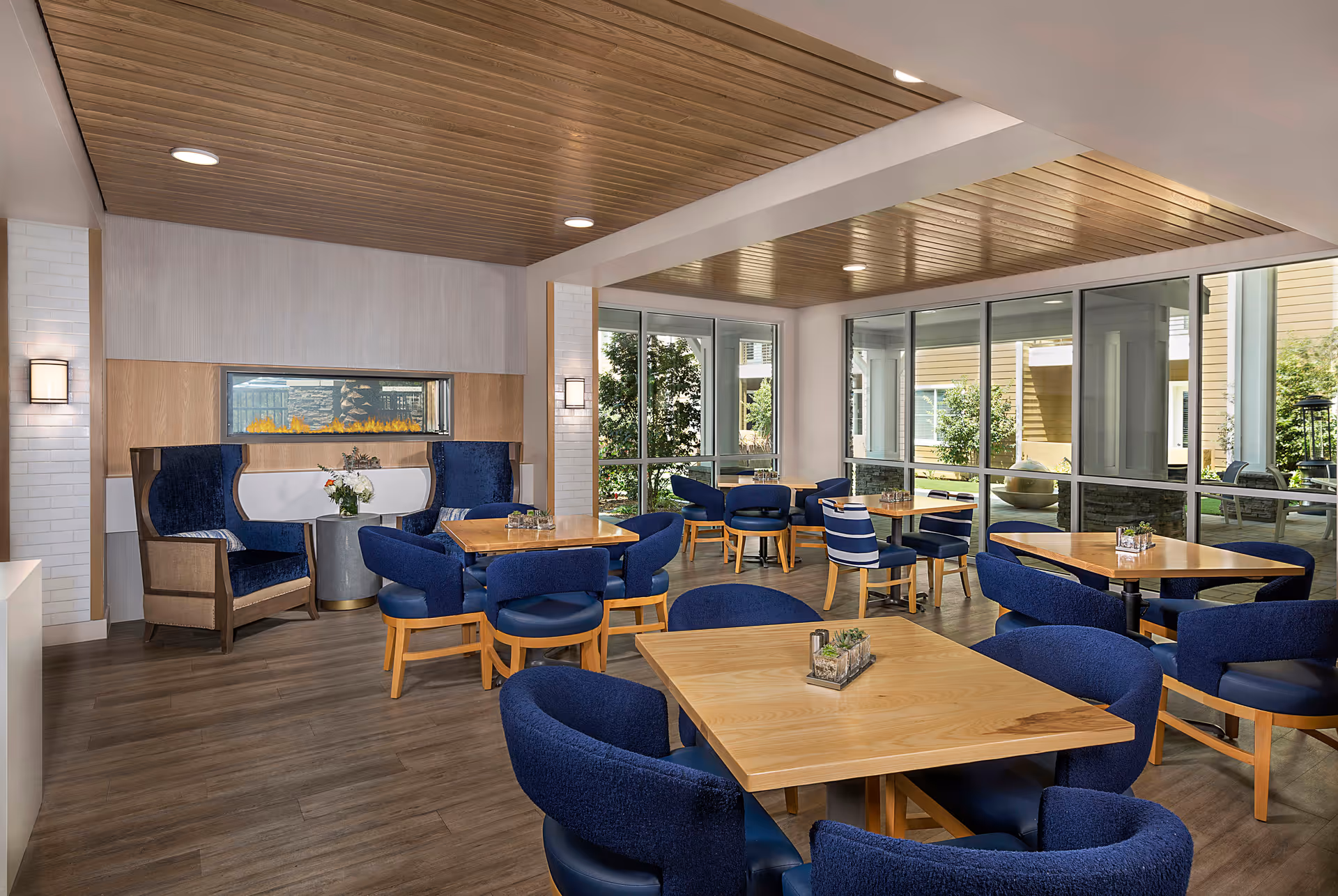 Communal dining/lounge room with wooden tables, blue upholstered chairs, large windows and a wood-paneled ceiling.