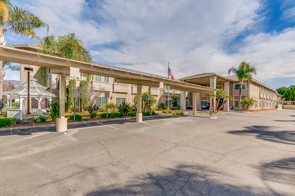 Front exterior of a two-story senior living building with a covered driveway, palm trees, a gazebo, and an American flag.