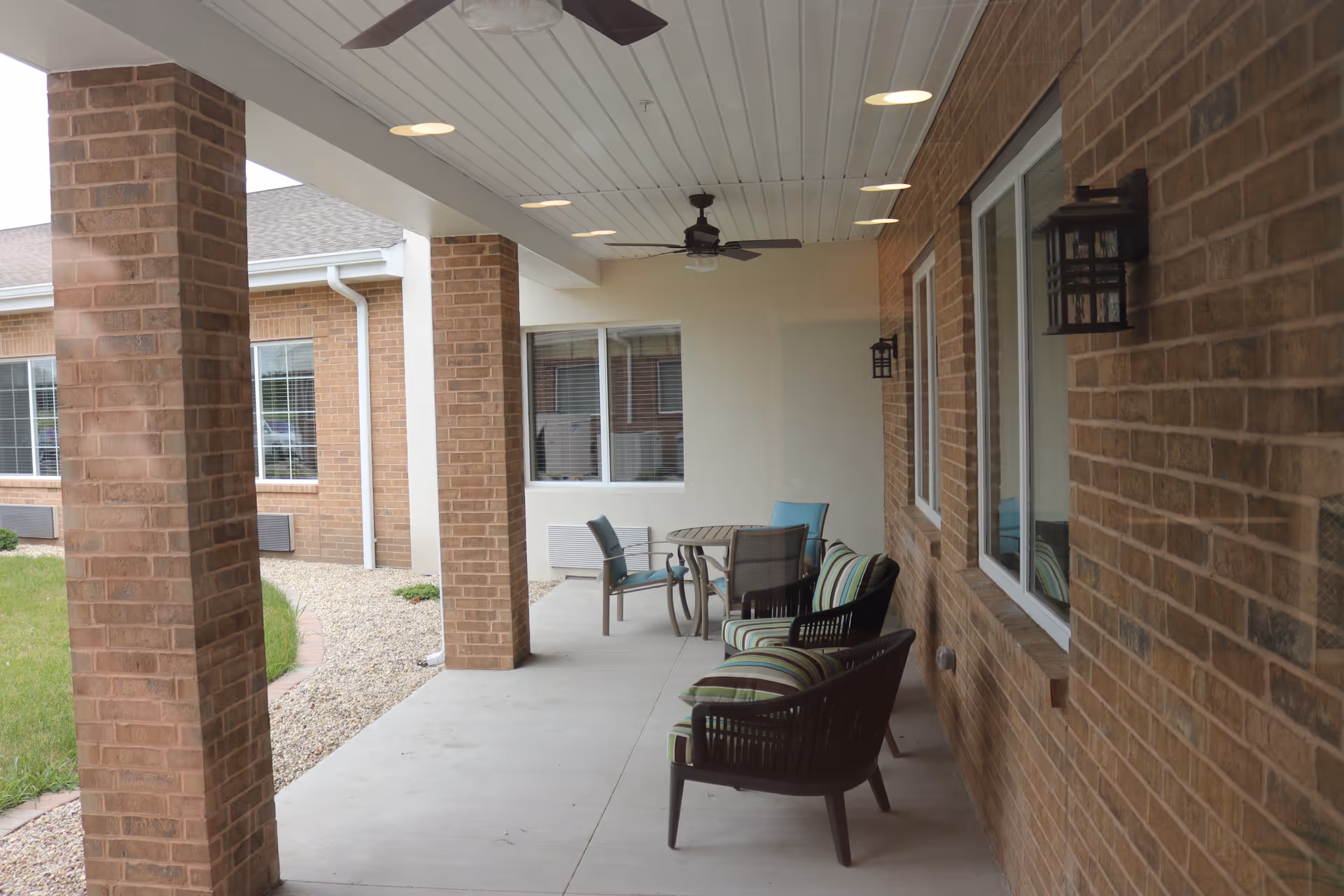 Covered outdoor patio area with brick pillars and walls, featuring ceiling fans, wall-mounted lantern lights, cushioned chairs, and a round table with chairs. The patio overlooks a small landscaped area with grass and gravel.