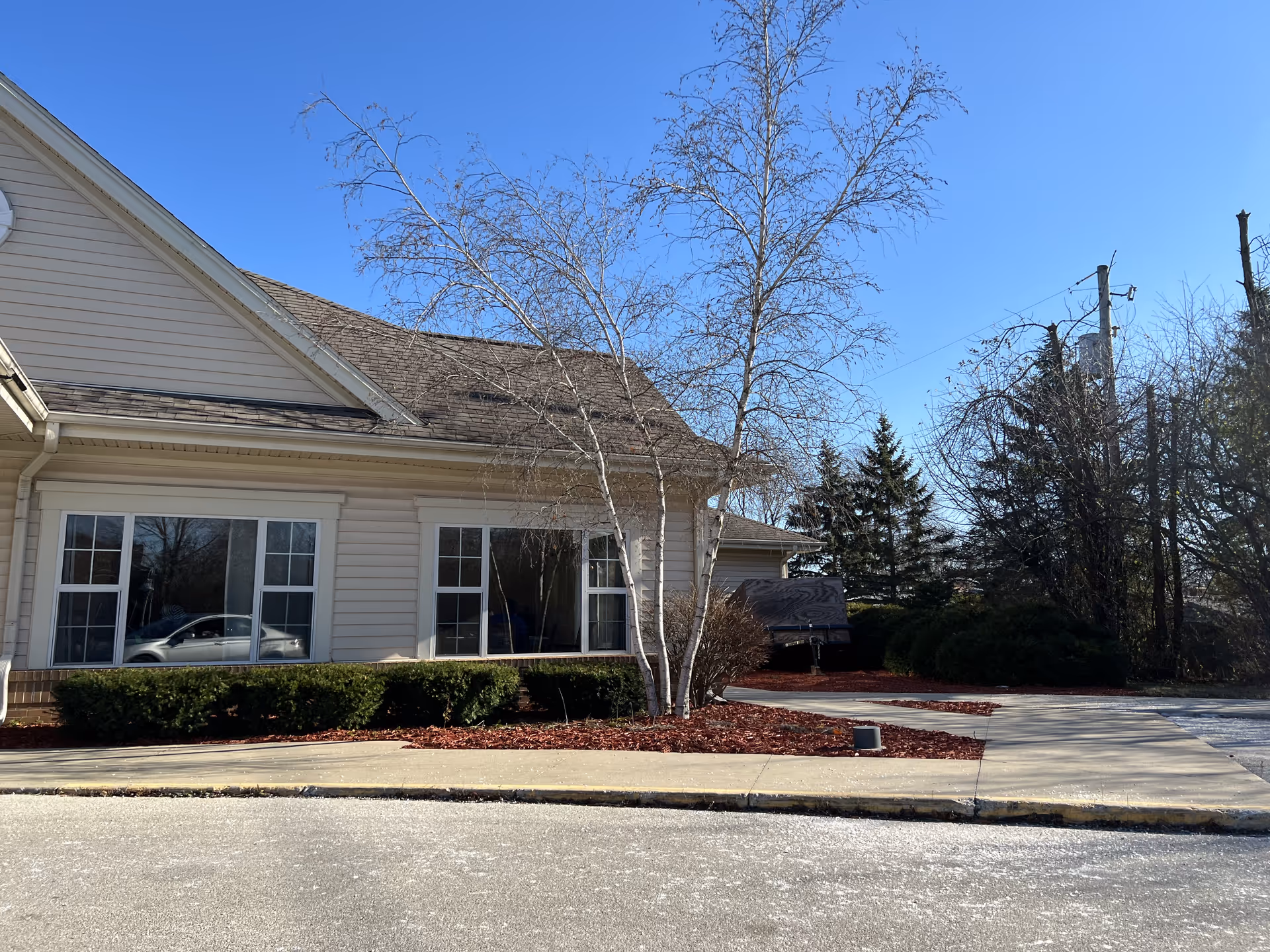 Exterior view of a beige building with large windows, a sidewalk, and landscaping including bushes and a leafless tree under a clear blue sky.