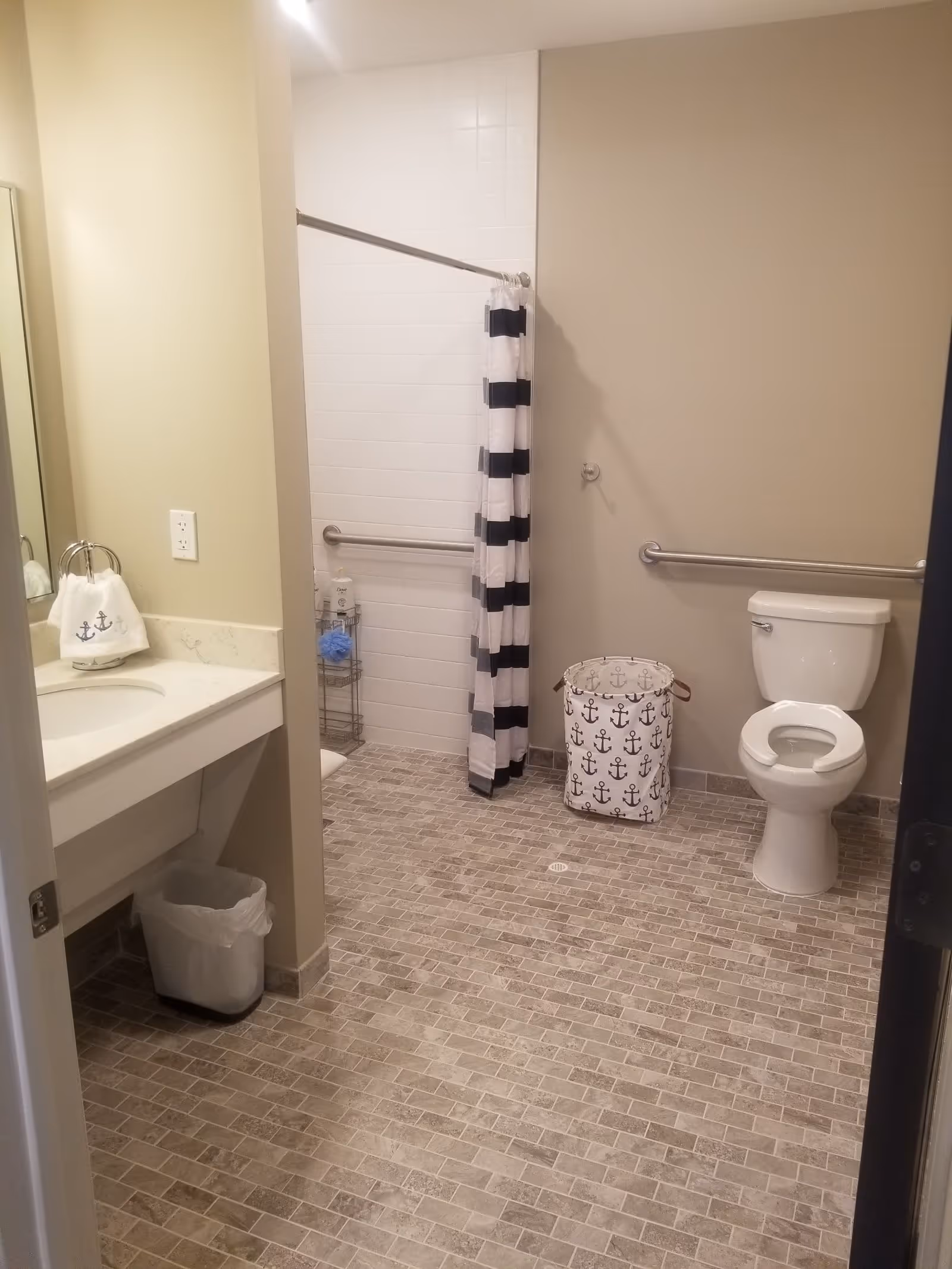 Accessible bathroom with a toilet, sink and mirror, grab bars, tiled floor, and a shower with a black-and-white striped curtain.